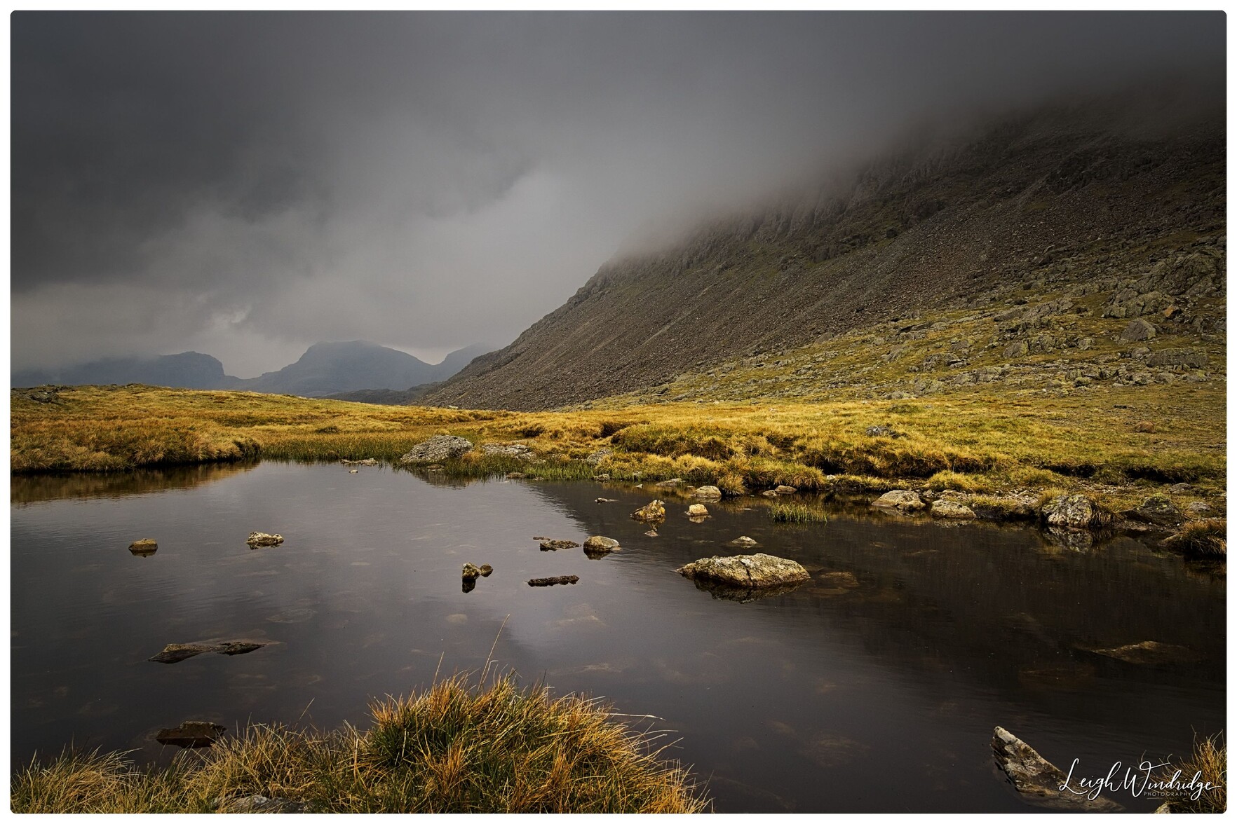 A view towards cloud strewn mountains with the cloud edging down to a large tarn of water in the foreground.