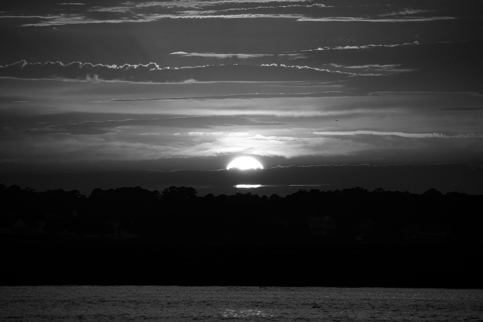 A black and white photo of a sunset over the Intracoastal Waterway in NC. Numerous horizontal clouds reflect the setting sun, including one that is bisecting it just above a ridge of trees on the opposite side of the water. There is a faint reflection of the setting sunlight on the calm water.